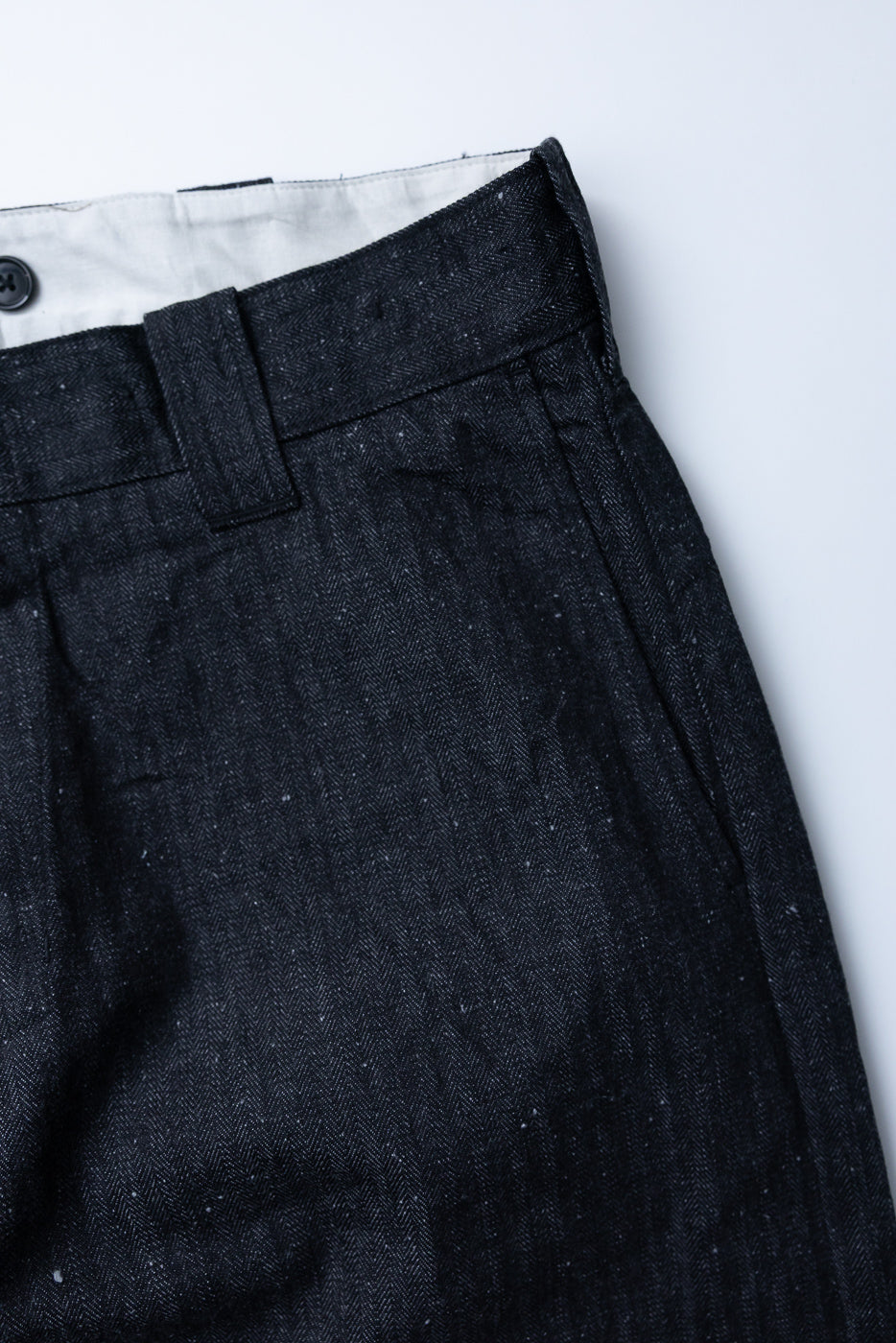 Close-up front view of dark herringbone wool trousers, showing waistband, button, and fabric texture