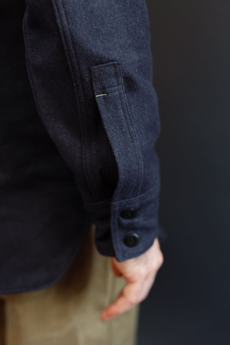 Close-up of navy shirt sleeve, showing button cuff and chest pocket with green stitching, worn by male model.