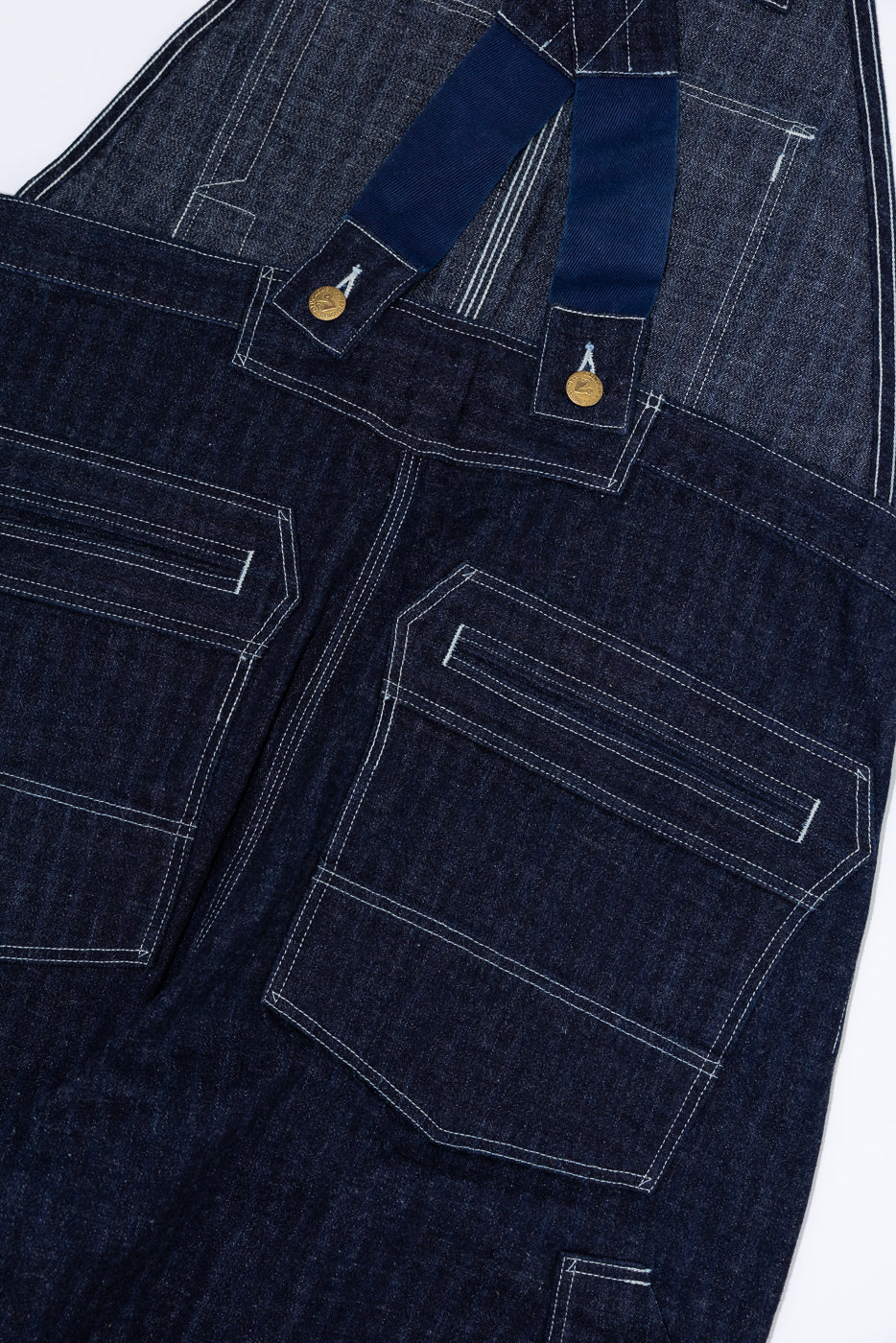Close-up front view of dark denim overalls with white stitching, gold buttons, and navy straps
