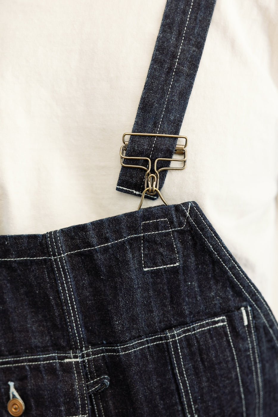 Close-up of dark denim overalls front, showing brass hardware and white stitching detail