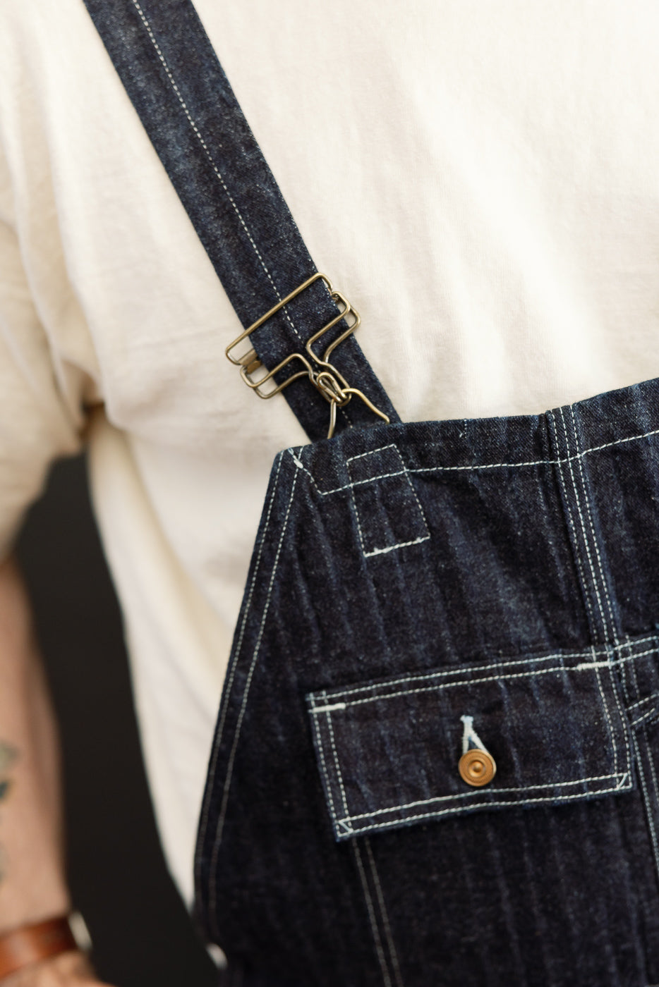 Close-up of male model's front denim overalls with brass buckle, button pocket, and white stitching.