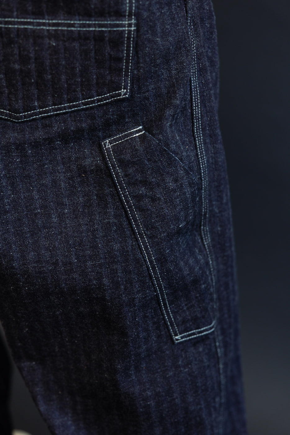 Close-up of dark selvedge denim jeans back pocket with contrast stitching, showing fabric texture and construction details.