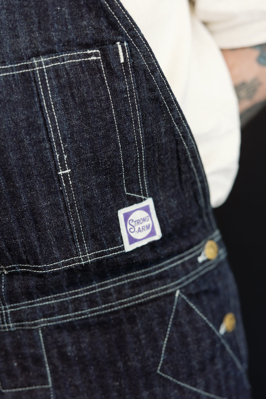 Close-up of dark selvedge denim overalls front pocket with white stitching, purple Strong Arm tag, and brass buttons
