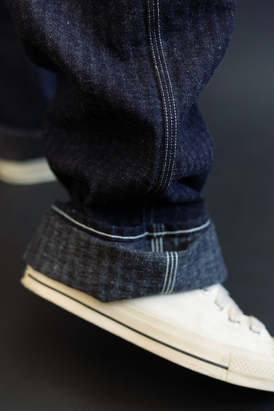 Close-up side view of dark selvedge denim jeans with cuffed hem, white contrast stitching, worn by male model with white sneakers