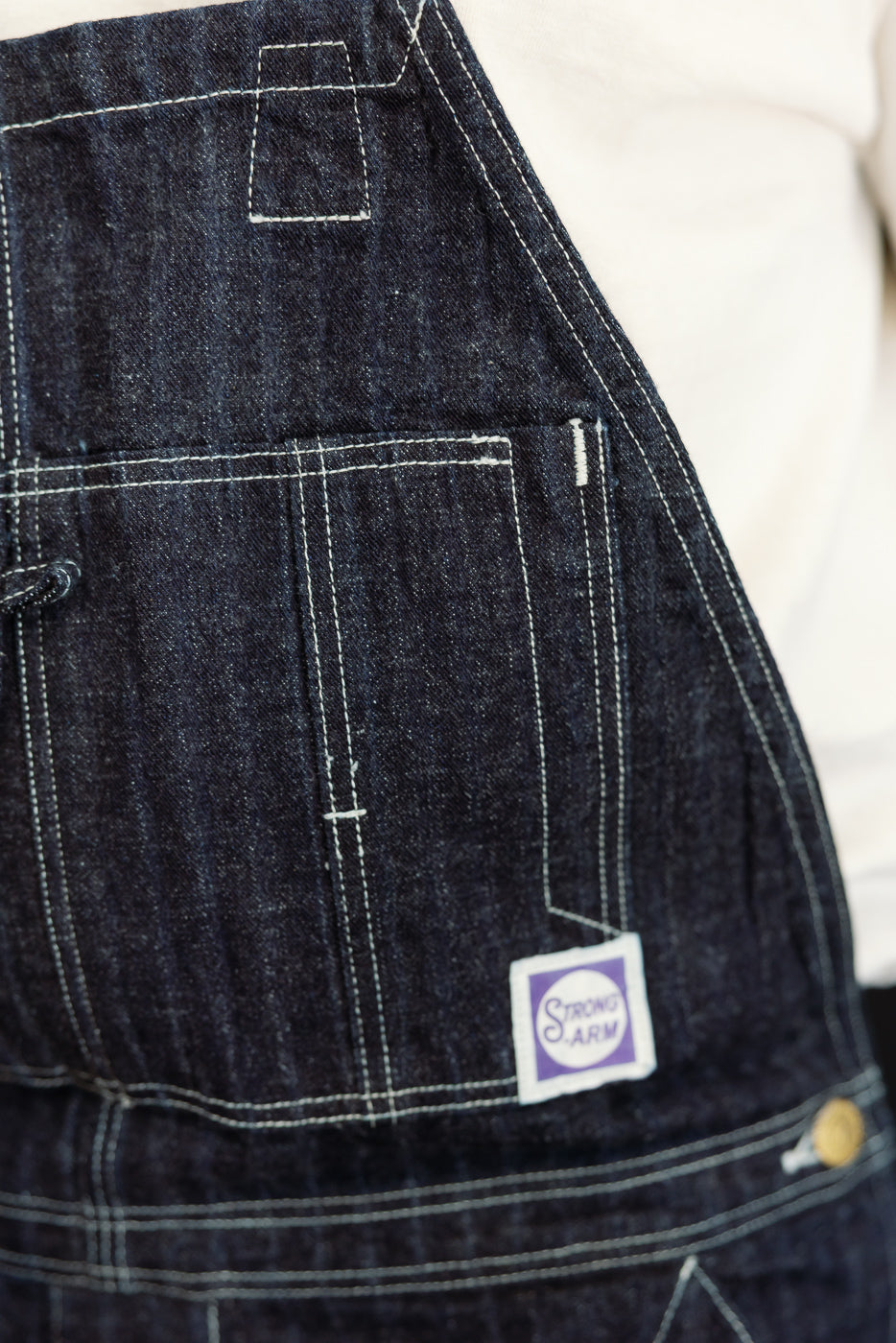 Close-up front right of dark selvedge denim overalls showing white stitching, pocket detail, and Strong Arm brand patch