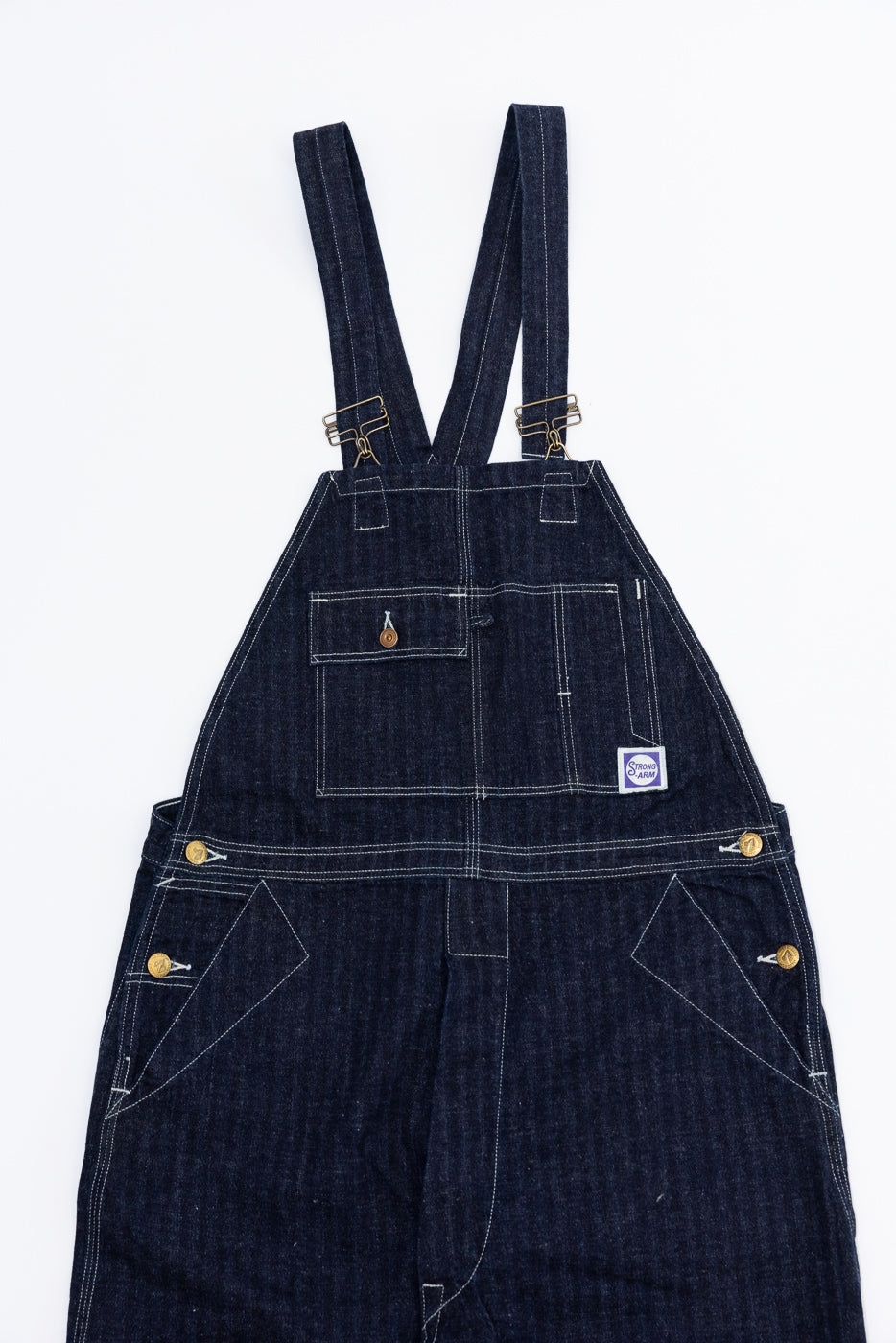 Front view of dark denim overalls with white stitching, brass buttons, and Strong Arm patch, flat lay