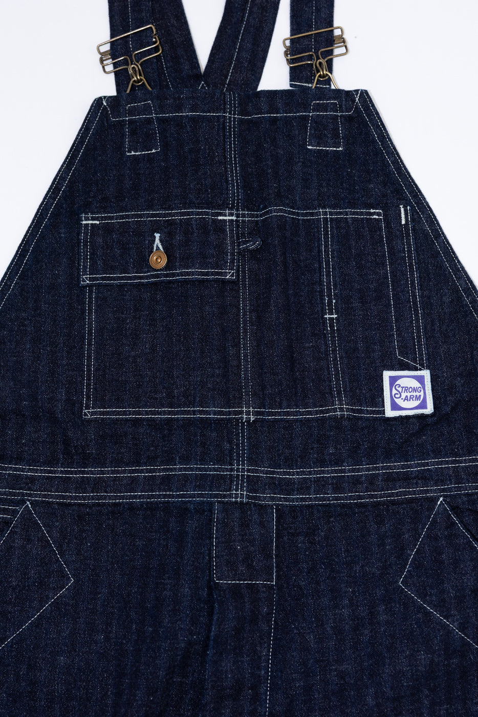 Front close-up of dark denim overalls with brass buttons, white stitching, and Strong Arm patch.