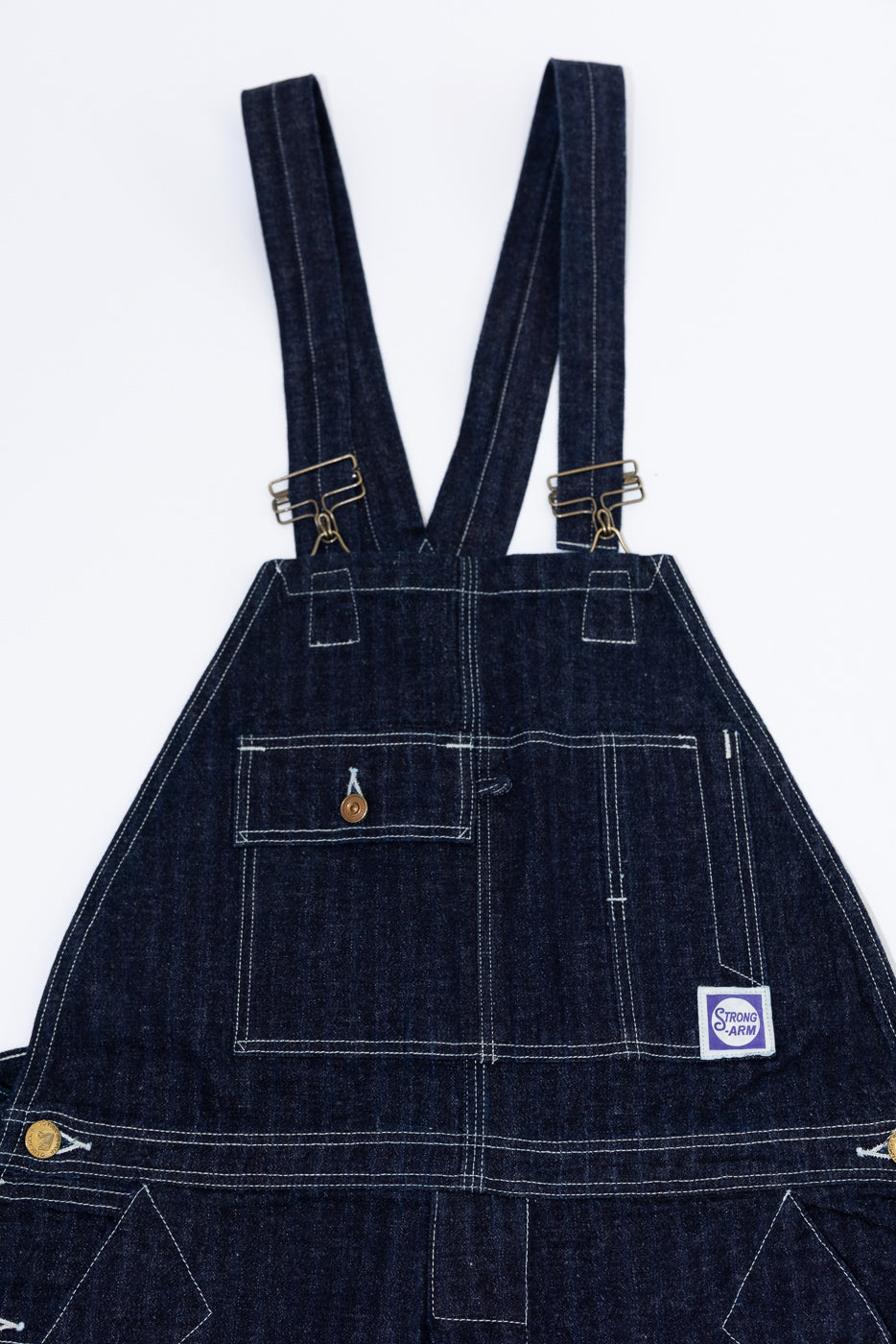 Front close-up of dark denim overalls with brass hardware, white stitching, chest pocket, and Strong Arm patch