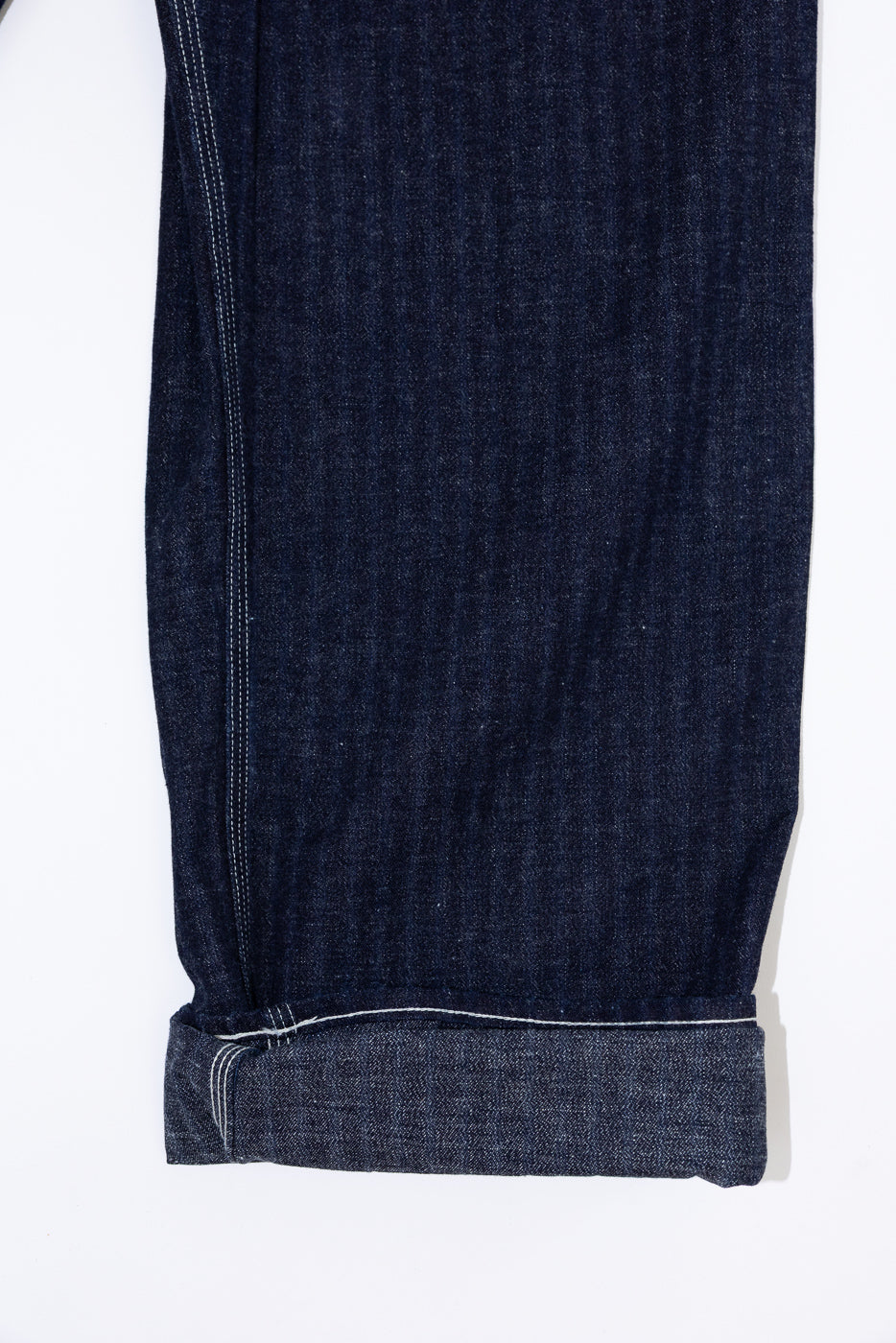 Close-up of dark selvedge denim jeans leg, front view, showing cuffed hem and white contrast stitching detail.