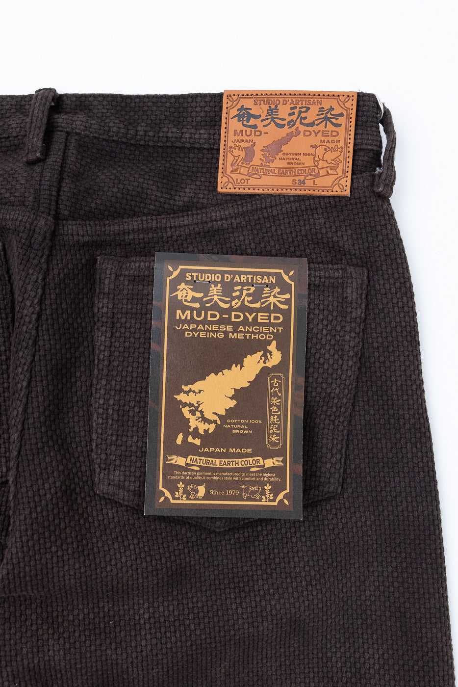 Close-up front view of mud-dyed Japanese selvedge pants, showing leather waist label and tag details, natural brown cotton texture.