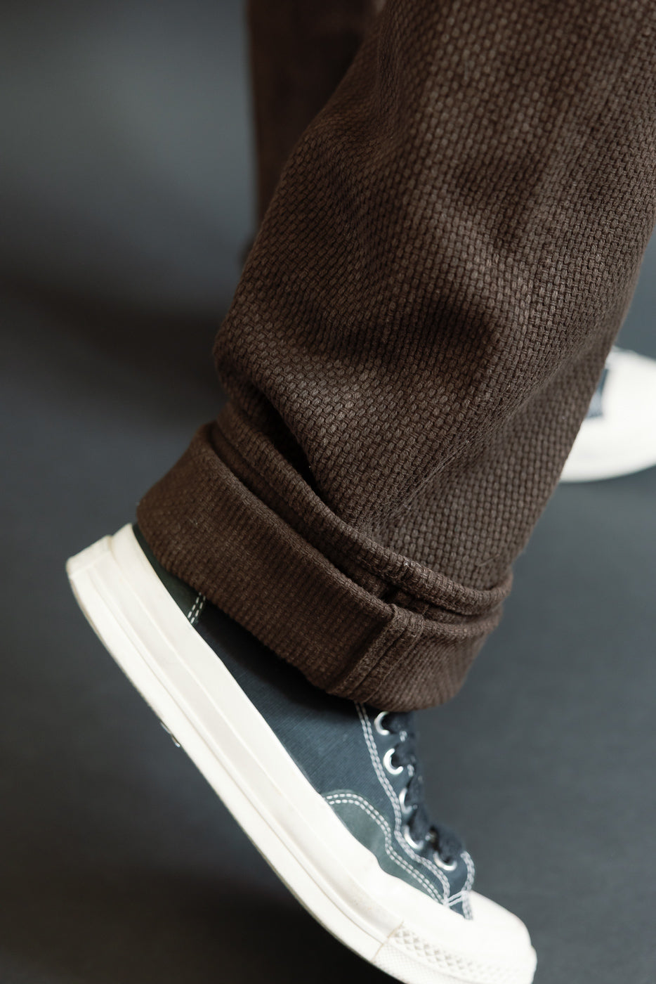 Close-up of male model's lower leg showing brown textured corduroy pants cuff over dark sneaker, front view