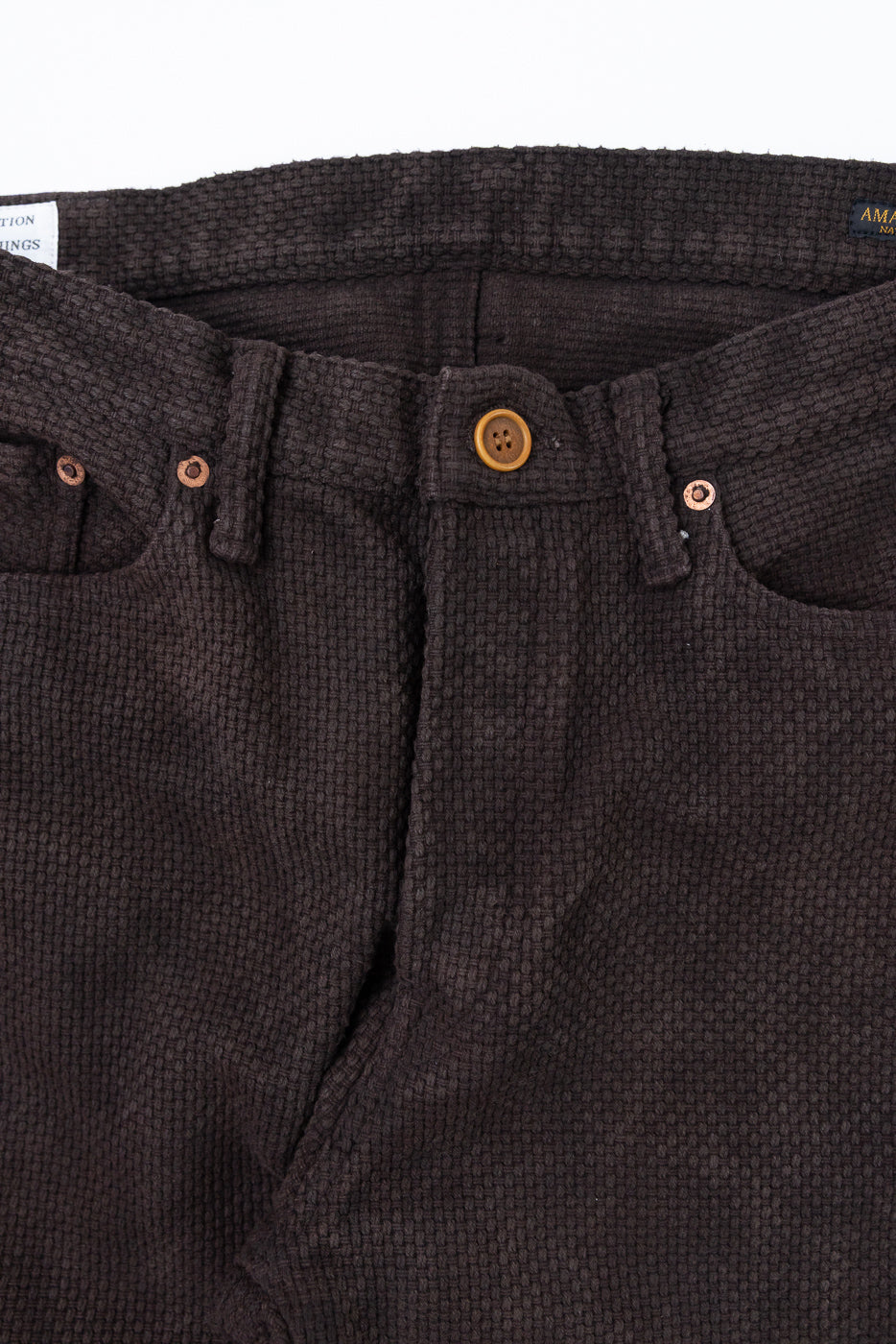 Close-up front view of dark brown textured pants with button fly and copper rivets