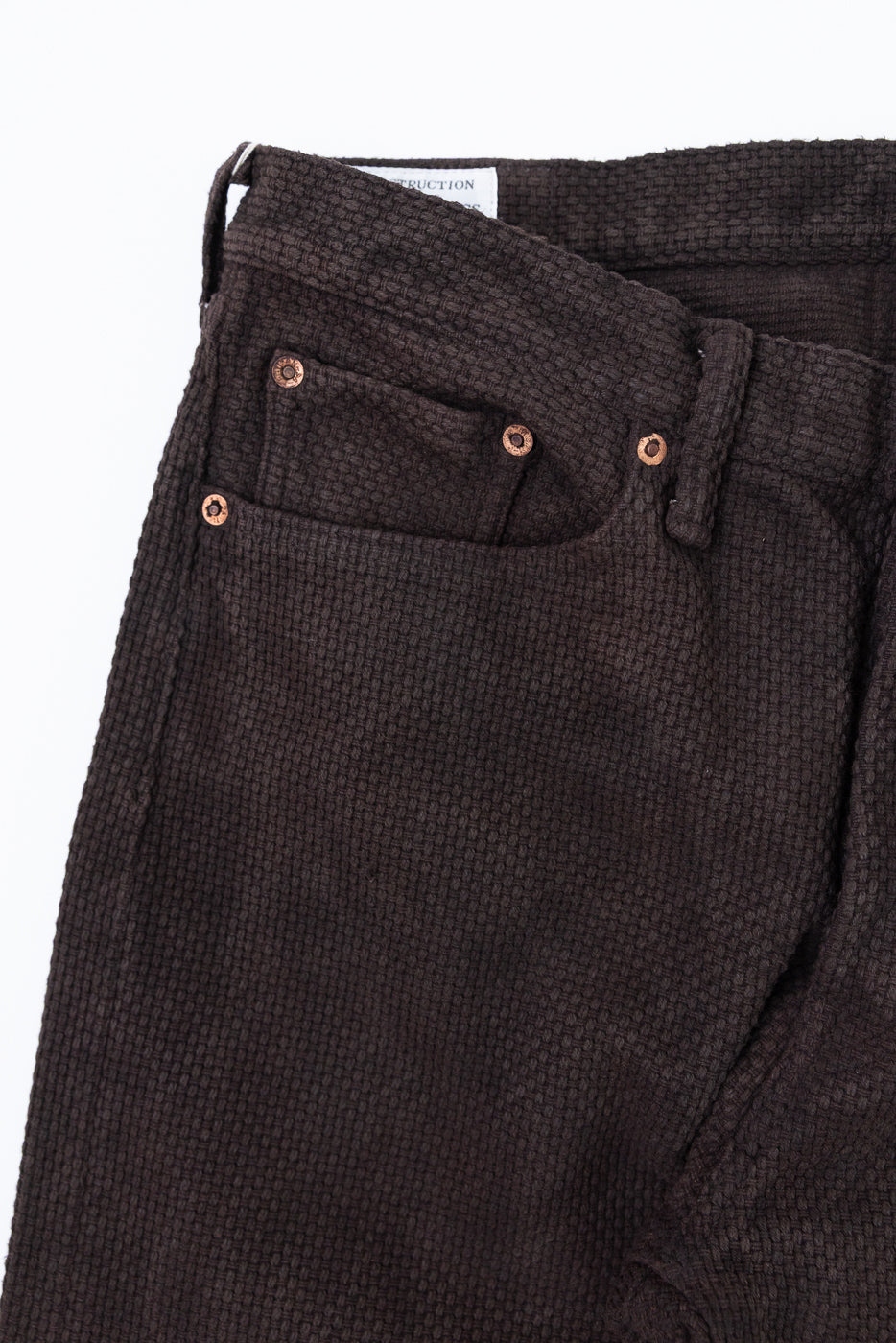 Close-up front view of dark brown textured pants with copper rivets and waistband tag
