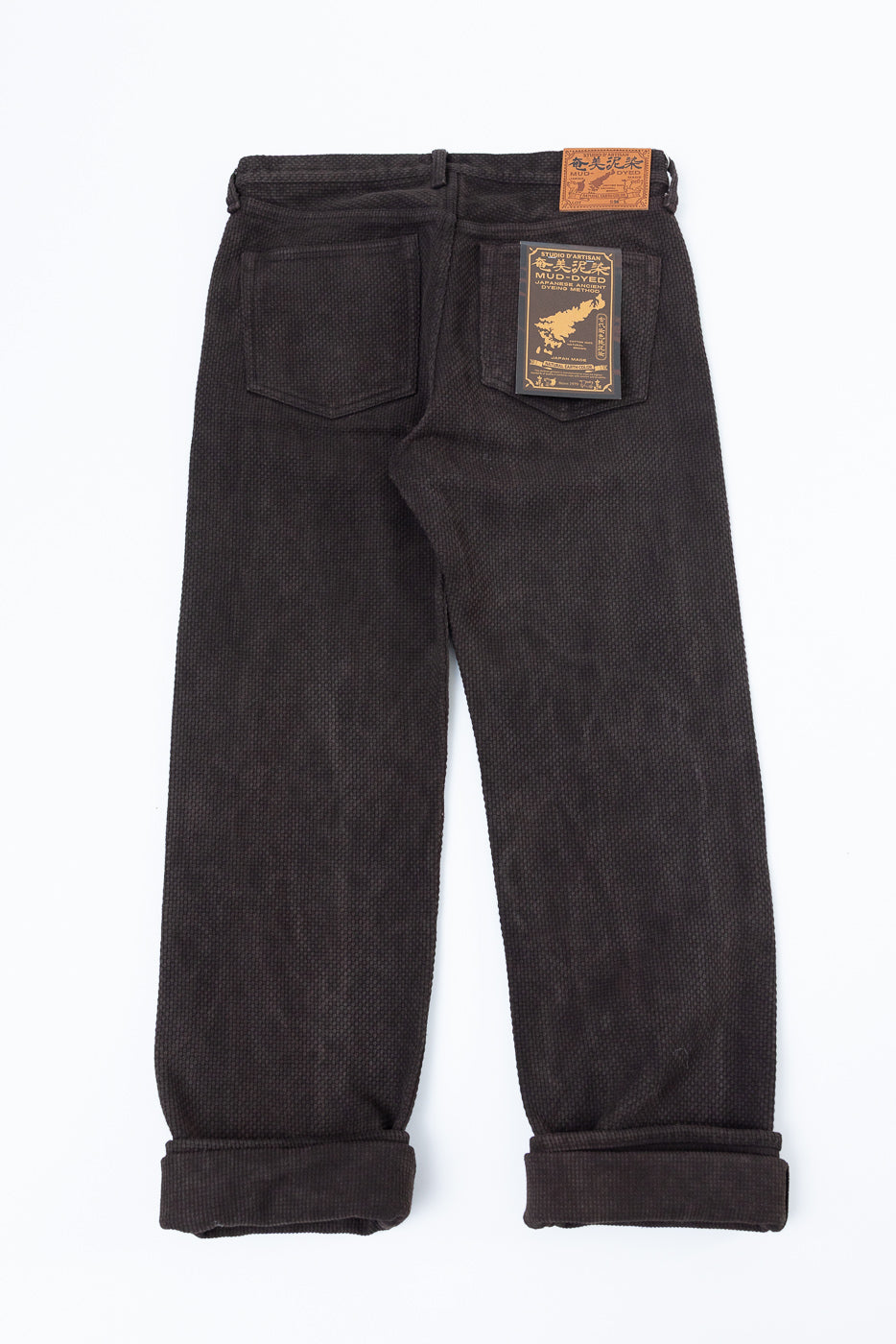 Back view flat lay of dark mud-dyed selvedge pants with cuffed hems, back pocket tag, and waist leather patch
