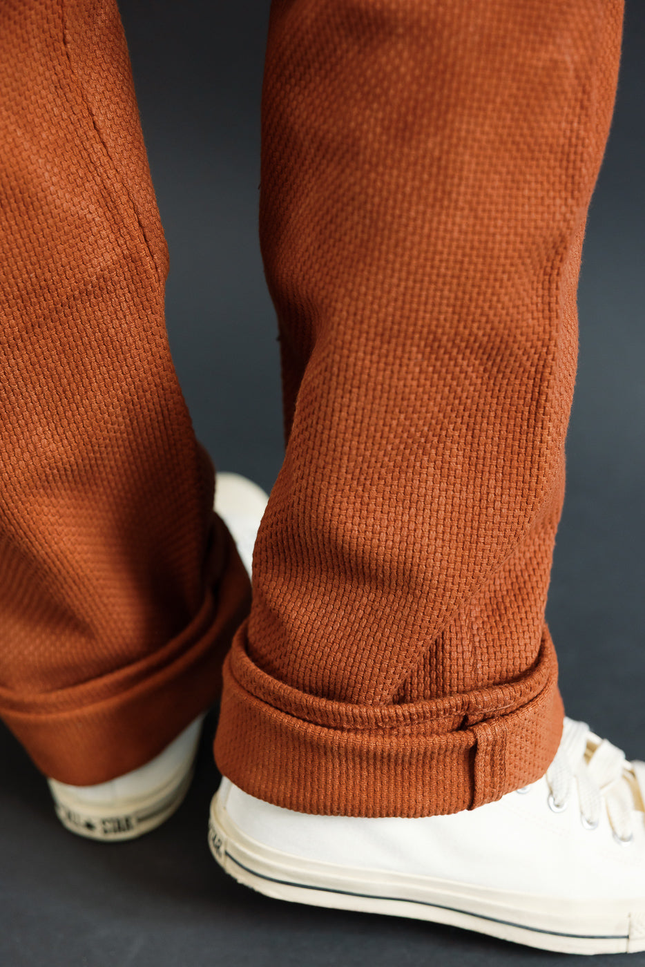 Close-up of male model's lower legs in rust corduroy pants with cuffed hems, paired with white sneakers