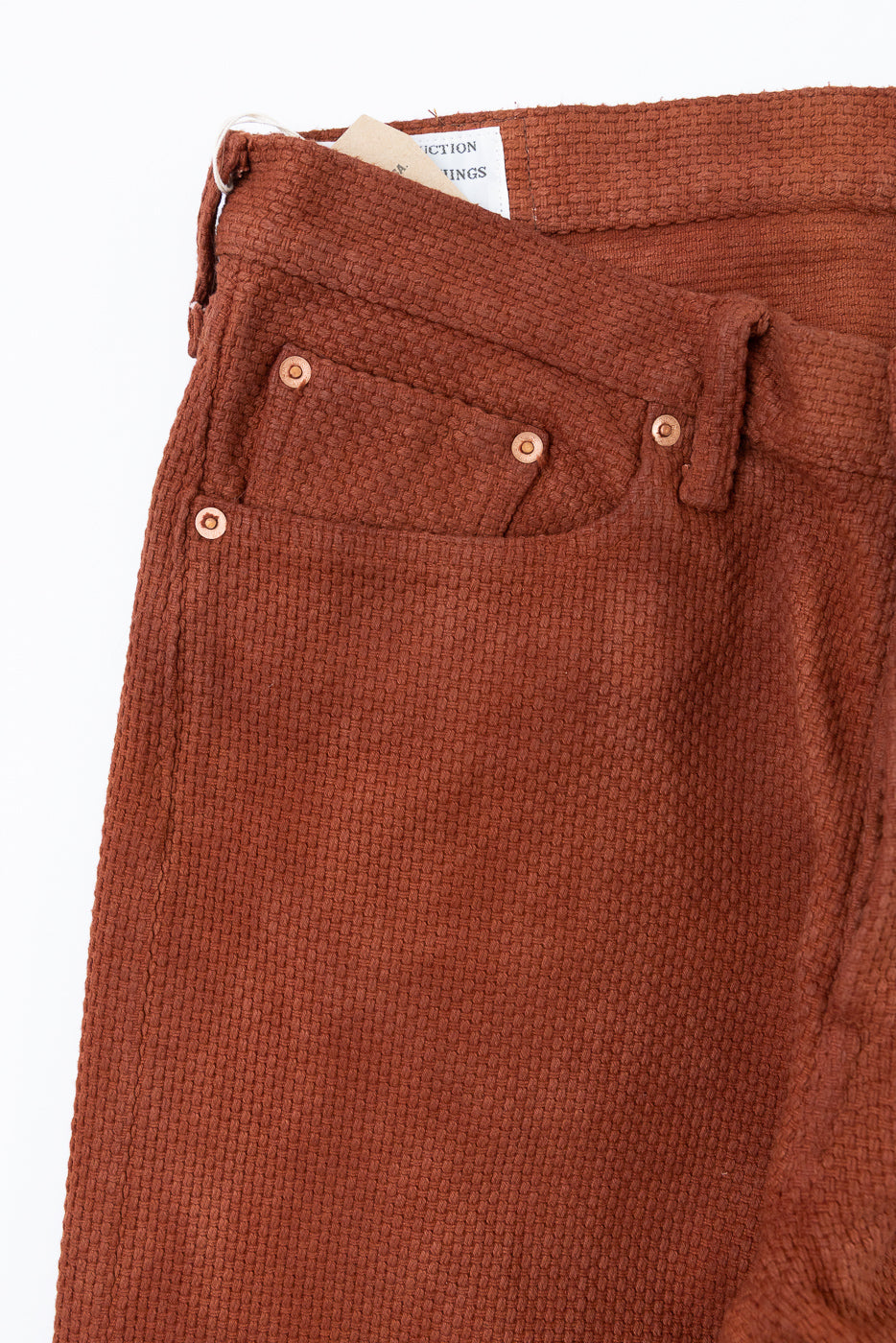 Close-up front view of rust-colored textured pants with copper rivets and waistband tag
