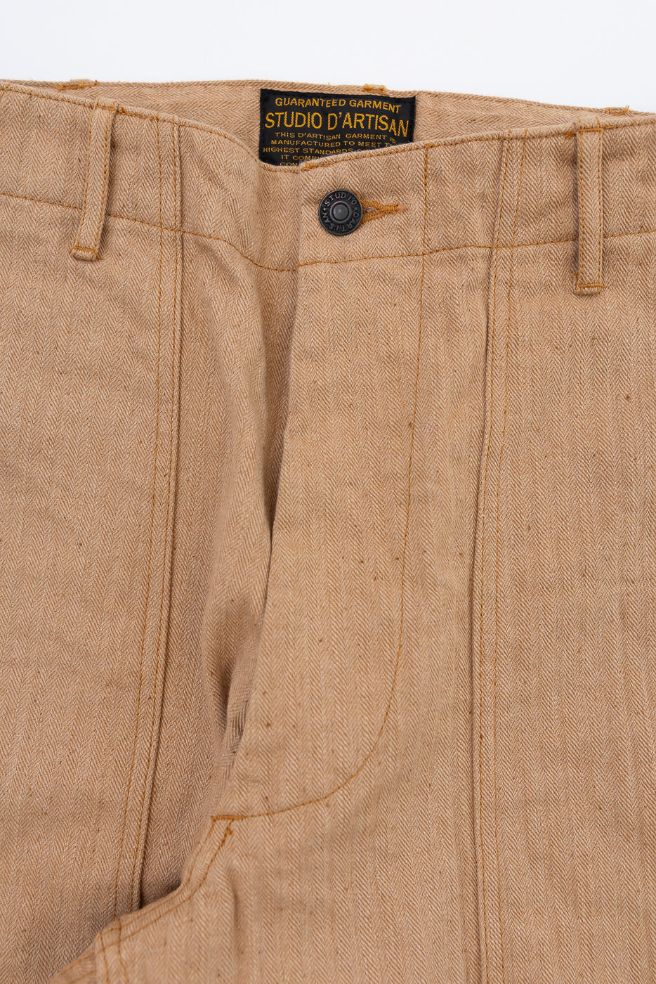 Close-up front waistband of tan herringbone pants, showing Studio D'Artisan tag, button, and stitching details