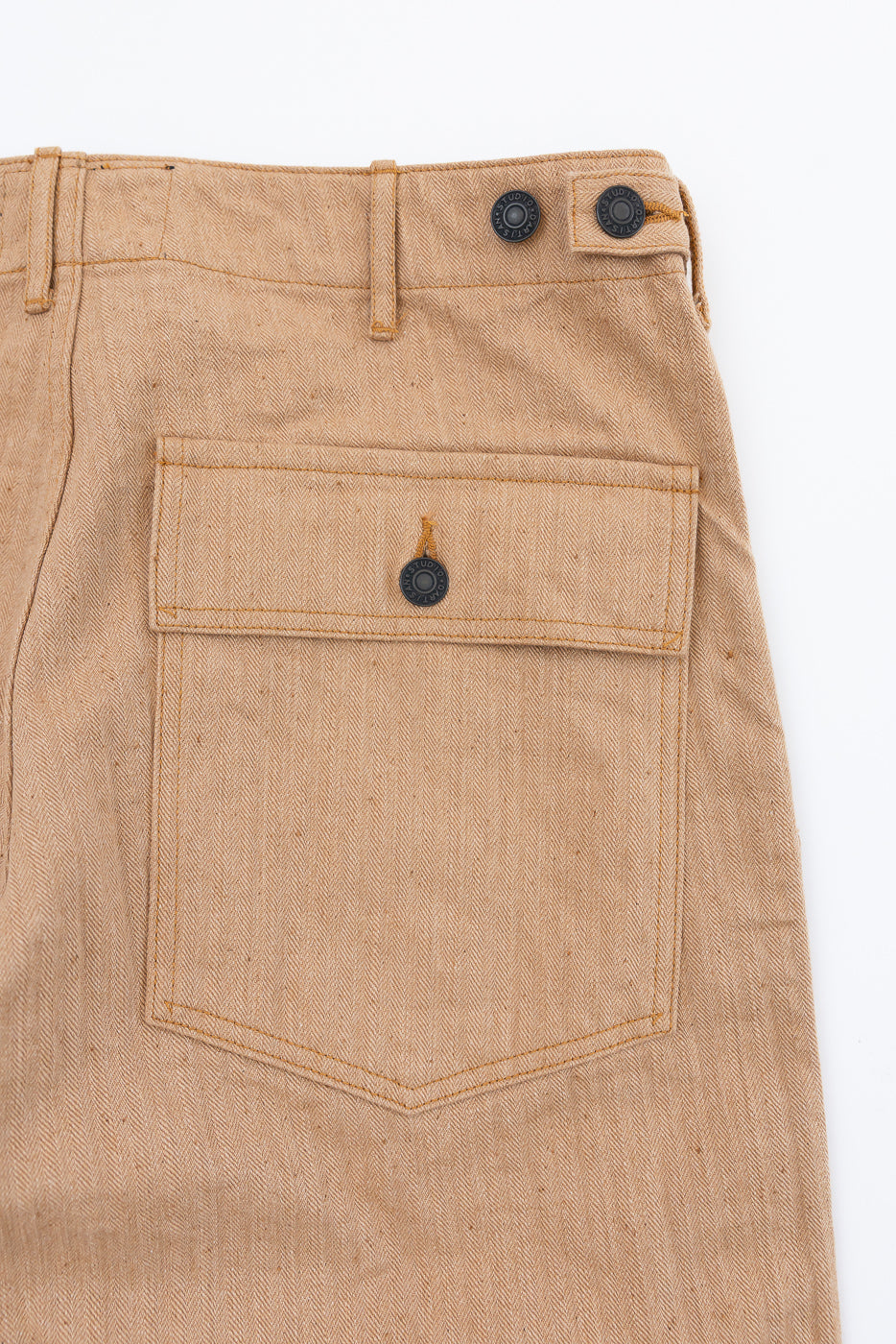 Close-up of beige herringbone pants front, showing buttoned pocket, waistband, and fabric texture