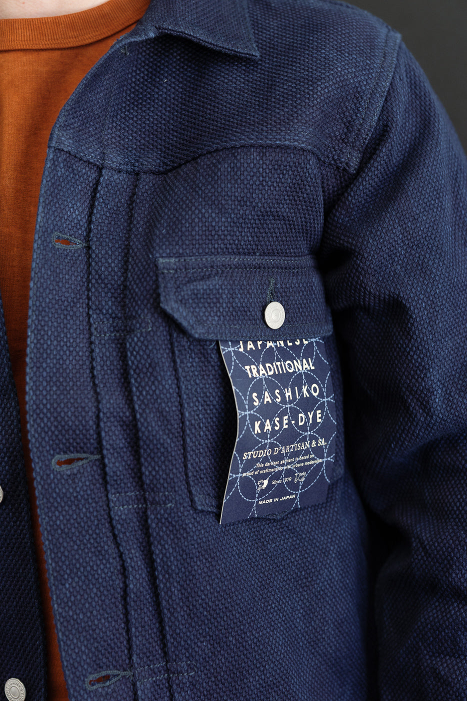 Close-up of navy jacket front, showing textured fabric, chest pocket, button, and Sashiko tag detail.