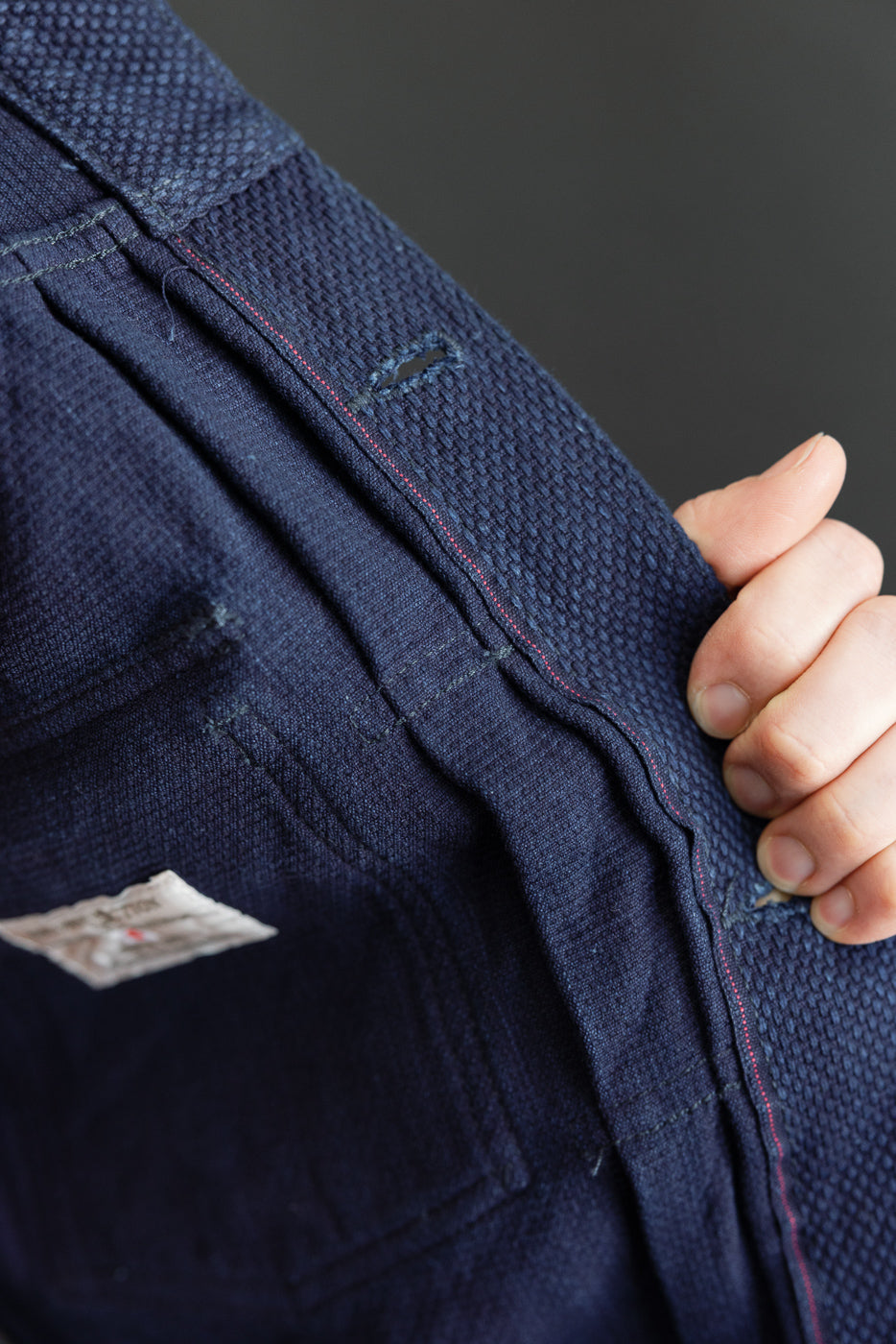 Close-up of dark indigo selvedge denim pants back pocket, showing red stitching, fabric texture, and woven tag detail.