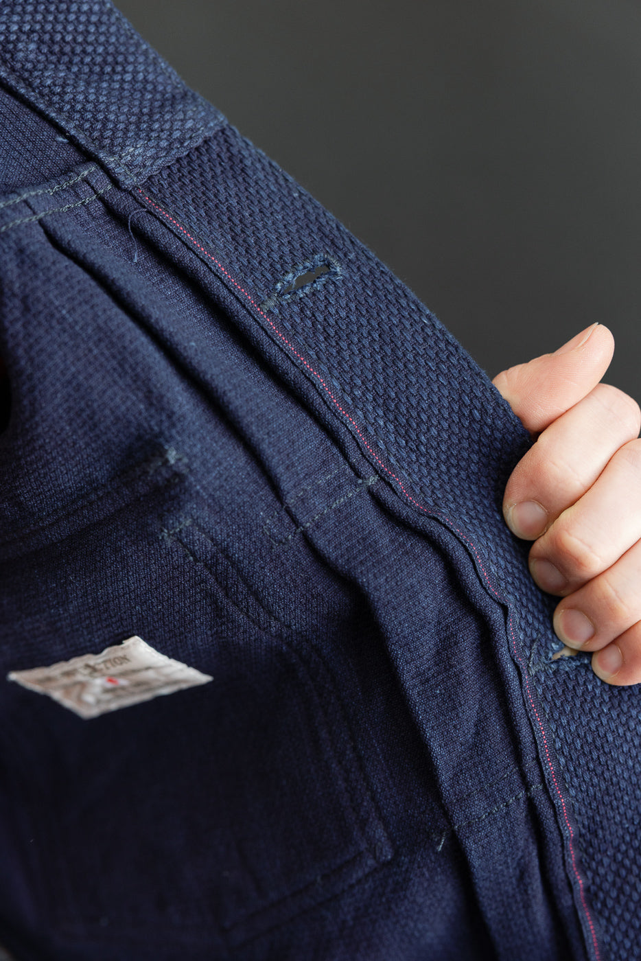 Close-up of dark blue selvedge denim pants, front view, showing red stitching, fabric texture, and tag detail
