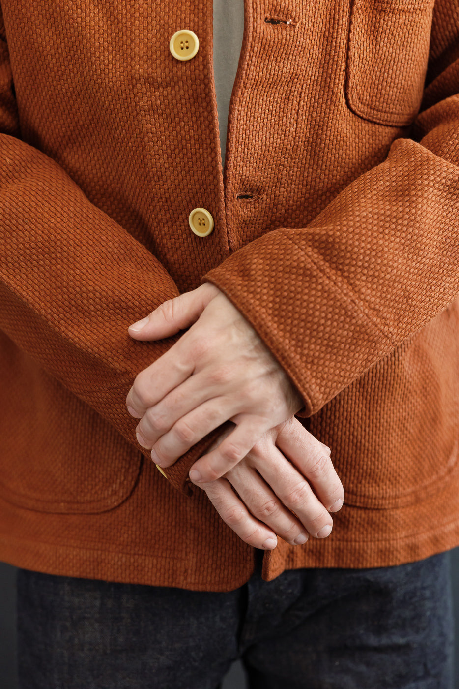 Close-up front view of a male model's hands and the textured fabric and buttons of an orange corduroy jacket.