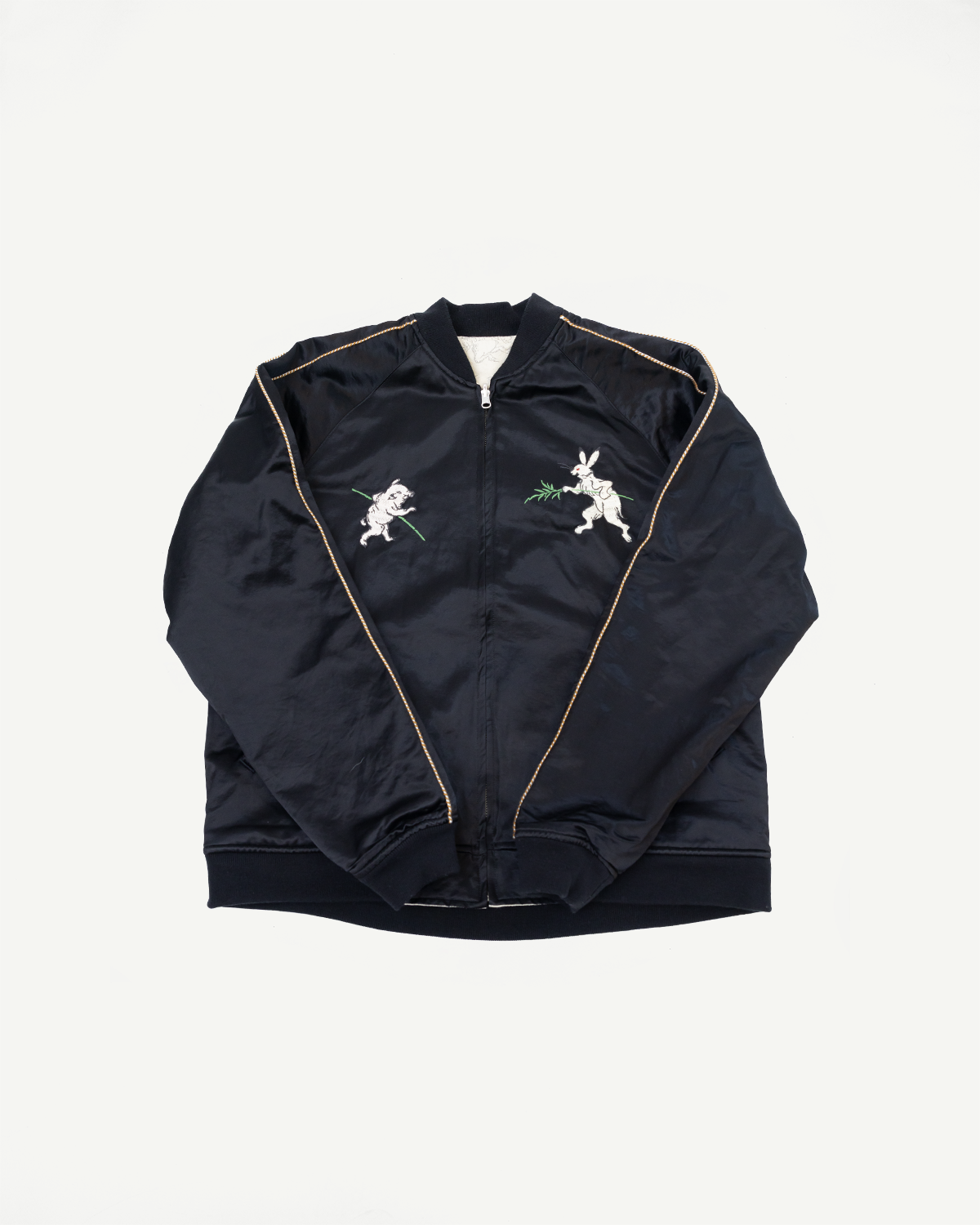 Black bomber jacket flat lay, front view, with rabbit embroidery and gold piping details.