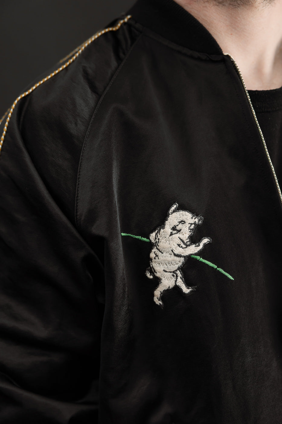 Close-up front view of black bomber jacket with embroidered bear on chest, gold piping, worn by male model