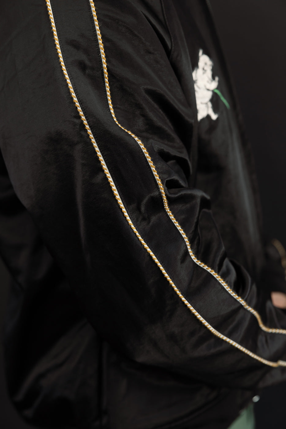 Close-up side view of black satin jacket with gold/white rope trim, white graphic on chest, worn by male model.