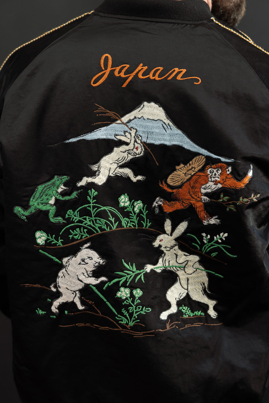 Back view close-up of black embroidered bomber jacket with Japan text, Mt. Fuji, rabbits, monkey, frog; male model 5'9