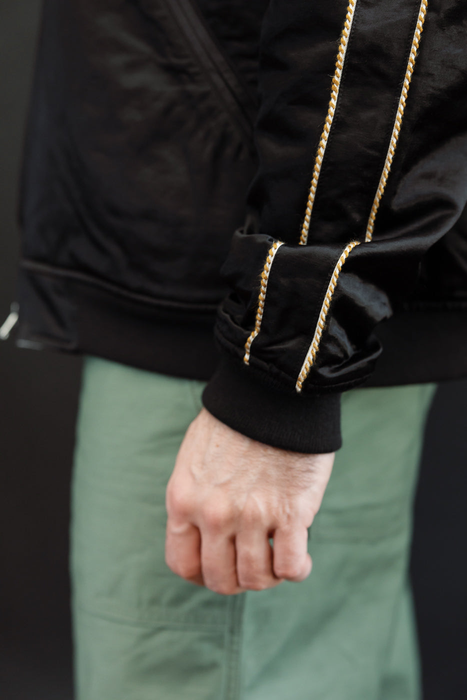 Close-up of male model's left sleeve: black bomber jacket with gold rope trim, ribbed cuff, athletic build.