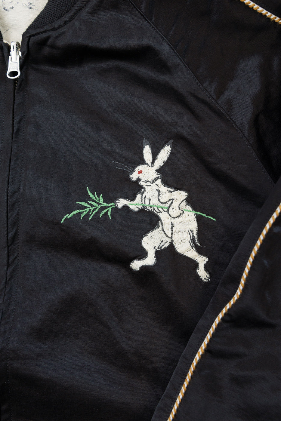 Close-up of black jacket front with embroidered rabbit holding green branch, gold stitching detail