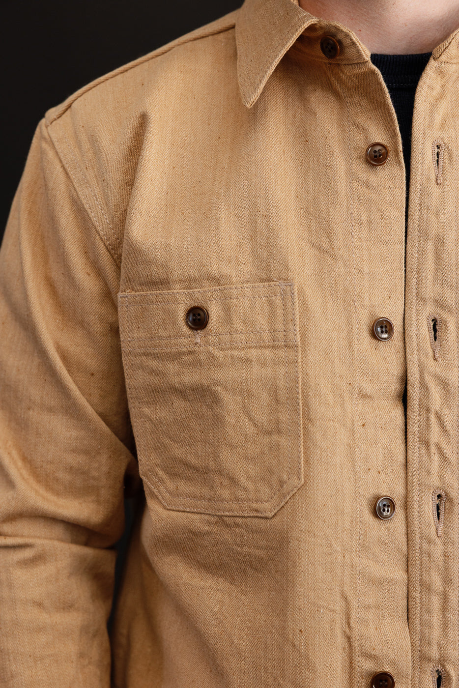 Close-up front view of male model wearing tan work shirt with button chest pocket, collar, and fabric texture