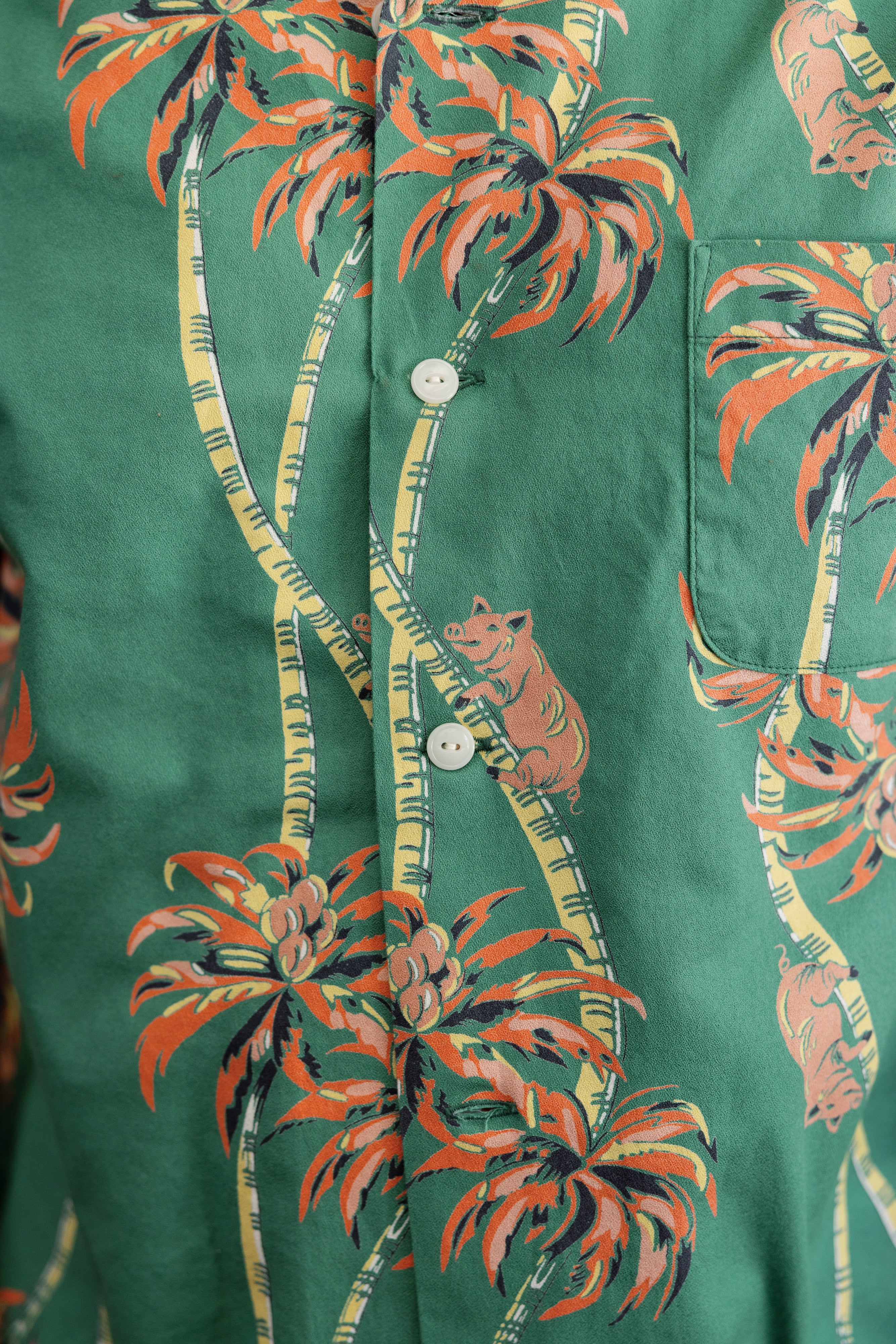 Close-up front view of green tropical print shirt with palm trees, pigs, white buttons, and chest pocket