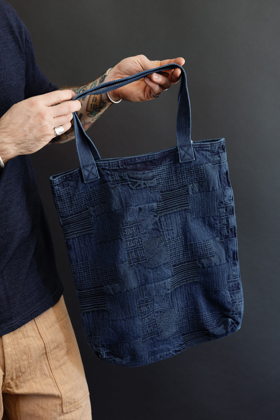 Male model holding indigo patchwork tote bag, front view, showcasing texture and handles