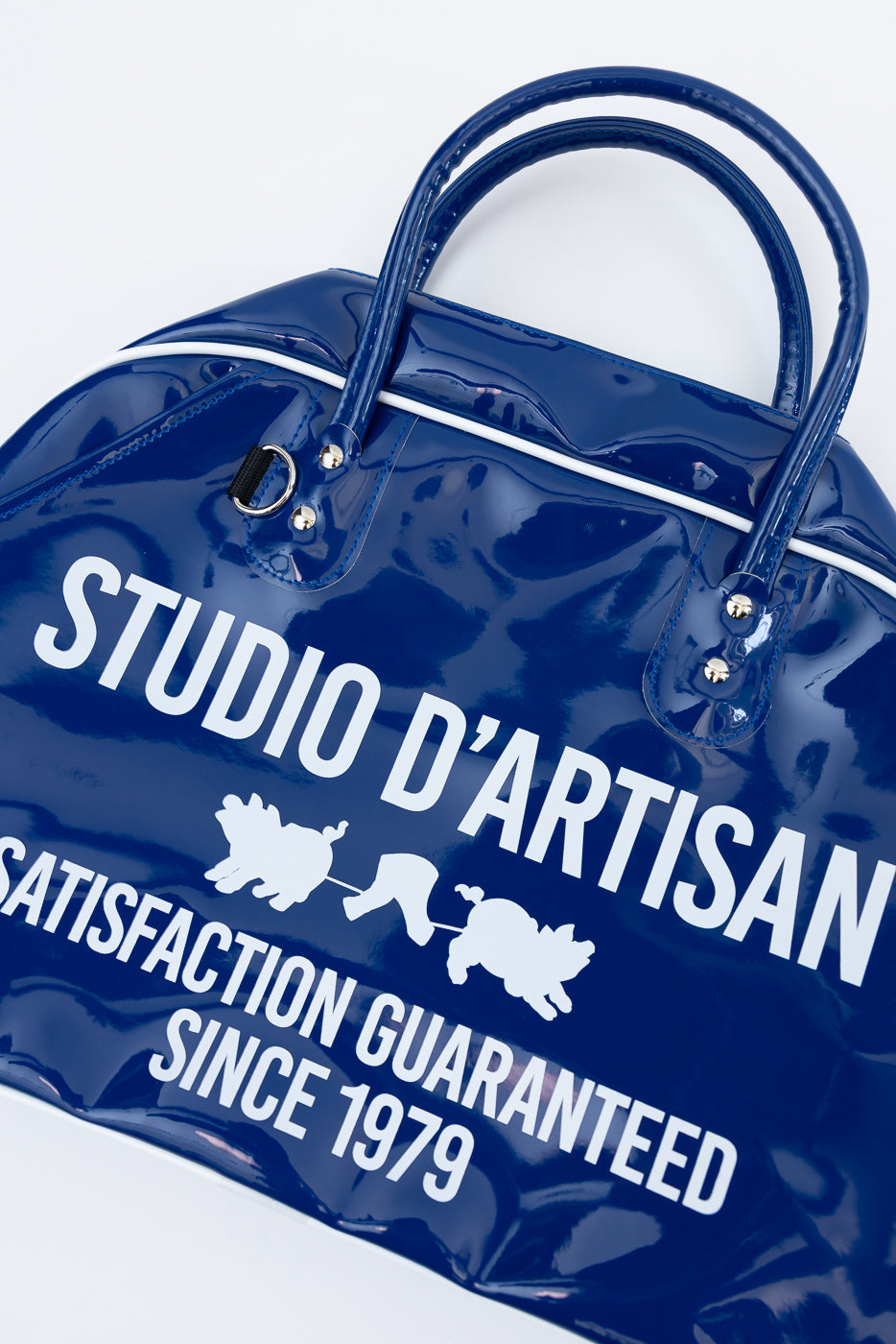 Close-up front of navy patent duffel bag with 'Studio D'Artisan' print, white stripe, and hardware details