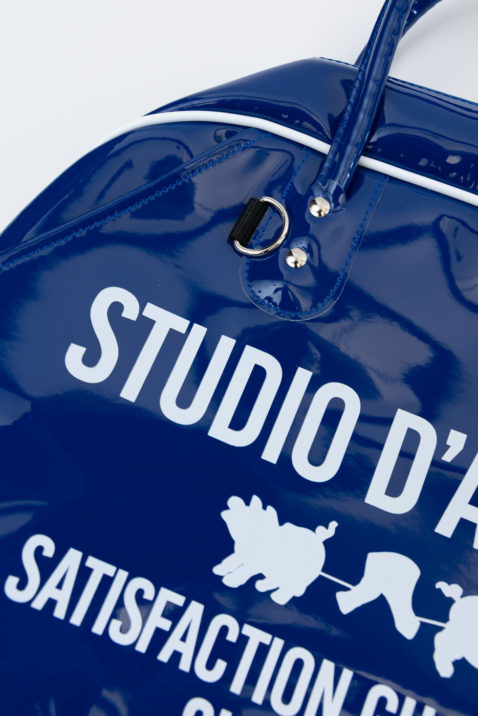 Close-up front view of glossy navy blue duffel bag with white 'STUDIO D' text, graphic print, silver hardware, and white trim.