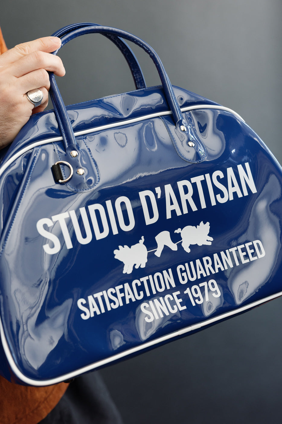 Close-up of blue glossy duffel bag front, 'STUDIO D'ARTISAN' text, white trim, metallic hardware, held by hand.