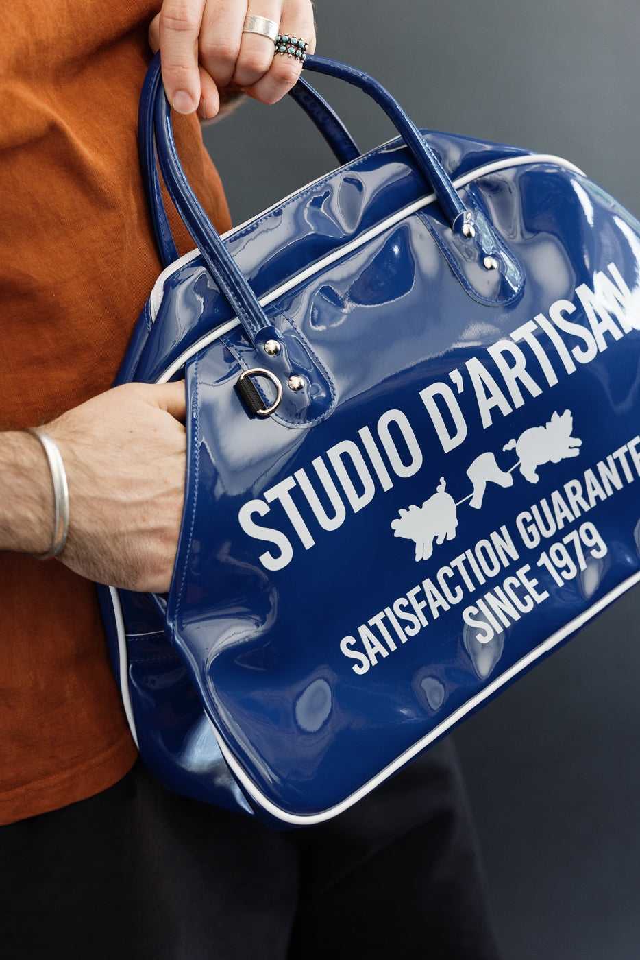 Close-up of male model holding glossy blue duffel bag, front view showing 'STUDIO D'ARTISAN' print, white trim, and hardware details.