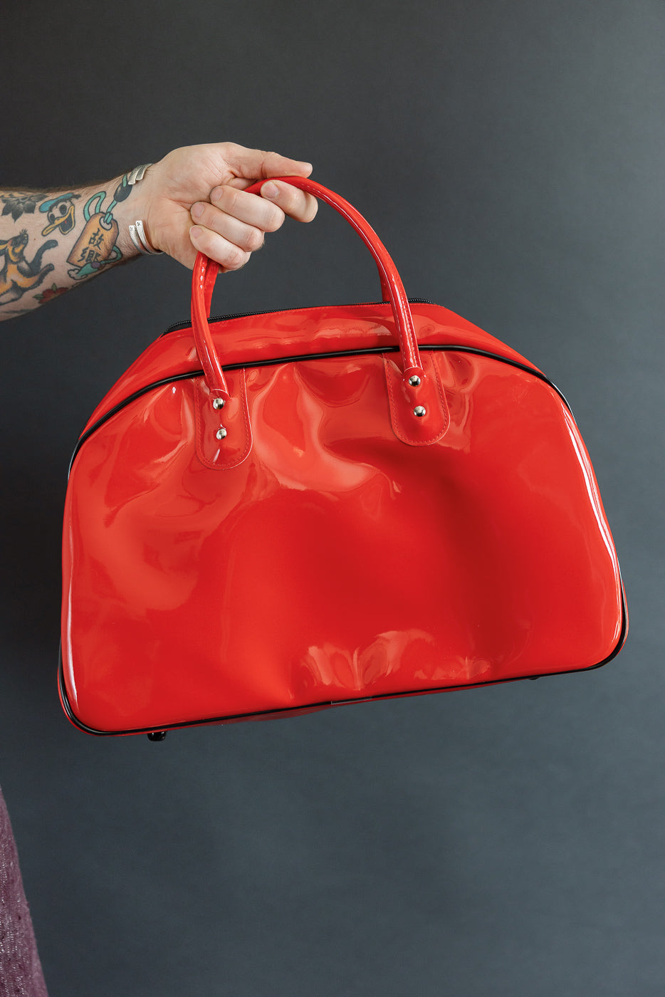 Red glossy handbag front view, held by tattooed hand, showcasing handles and silver hardware