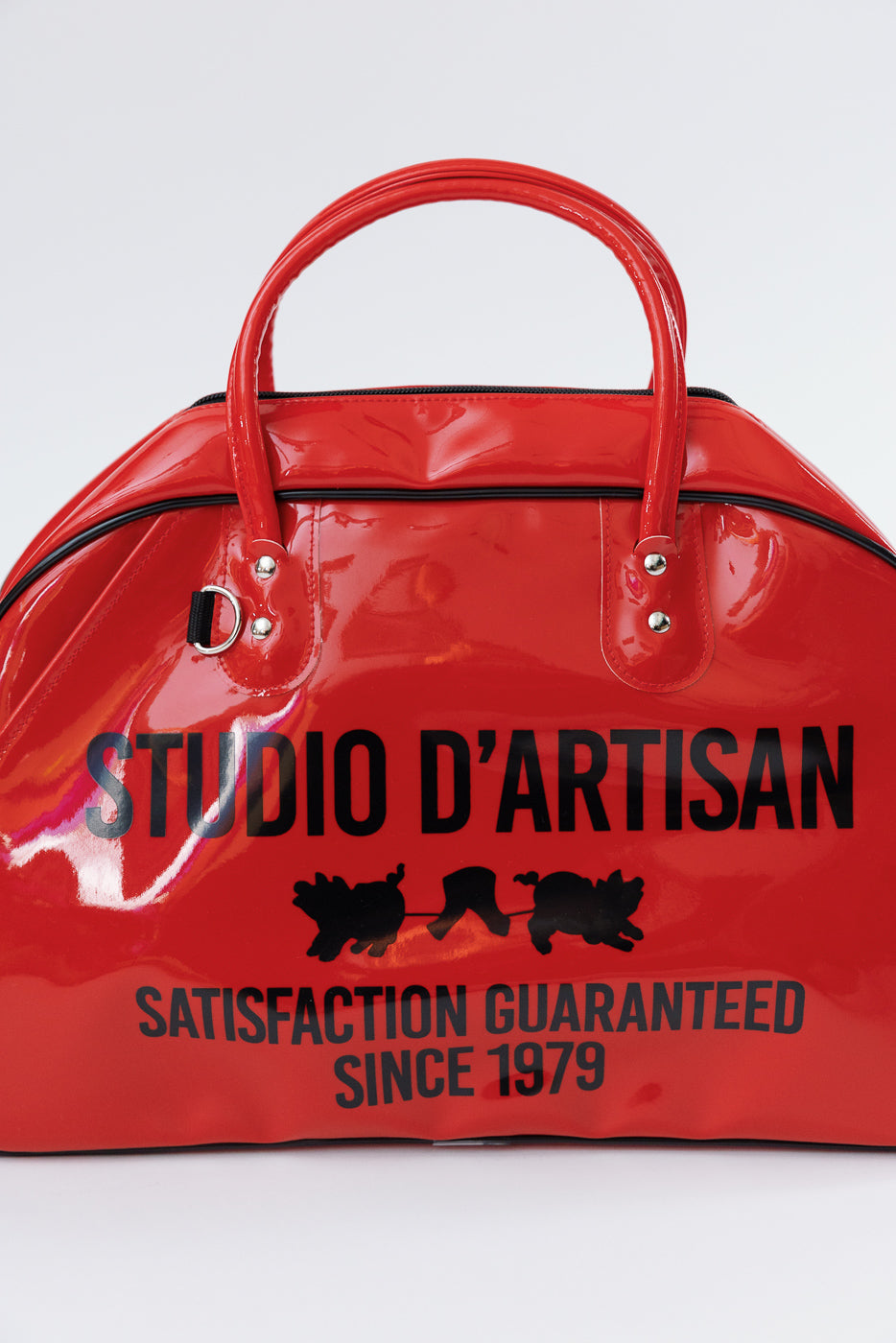 Red glossy duffel bag front view, 'STUDIO D'ARTISAN' print, black text, silver hardware, zip closure