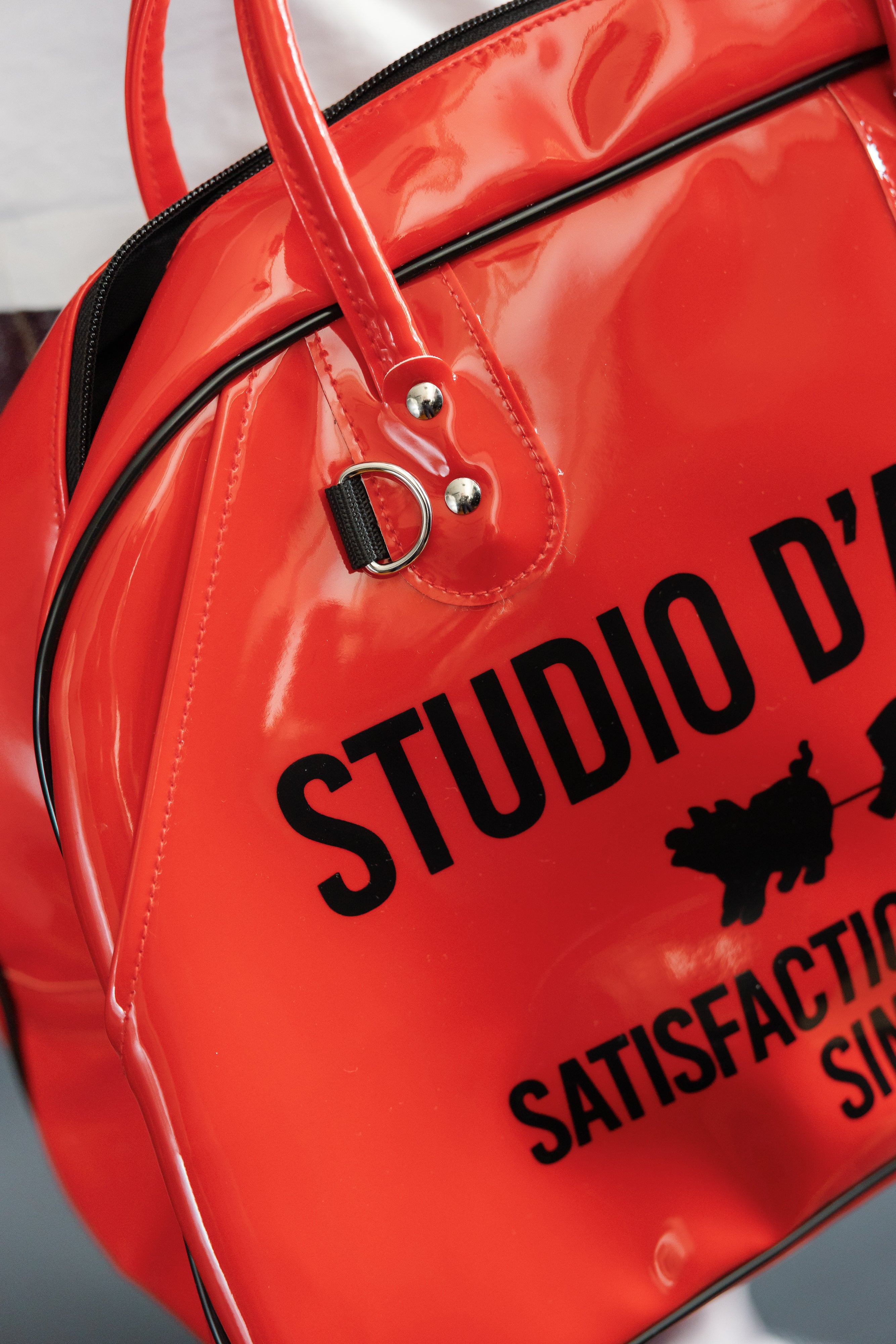 Close-up of red glossy bag front, showing 'STUDIO D' text, black trim, and silver hardware details.