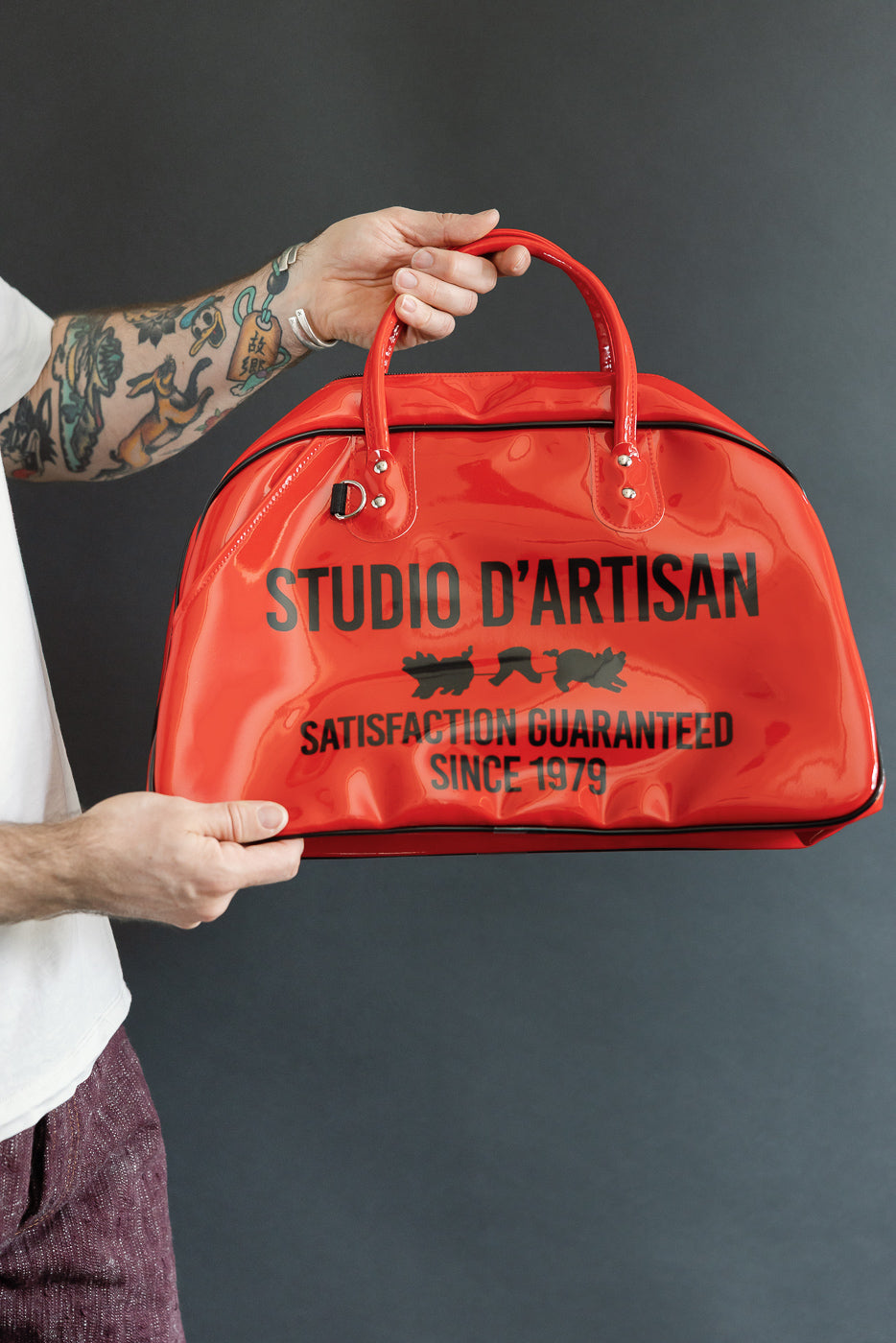 Male model holding red glossy duffel bag front view, 'STUDIO D’ARTISAN' print, black trim, 5'9