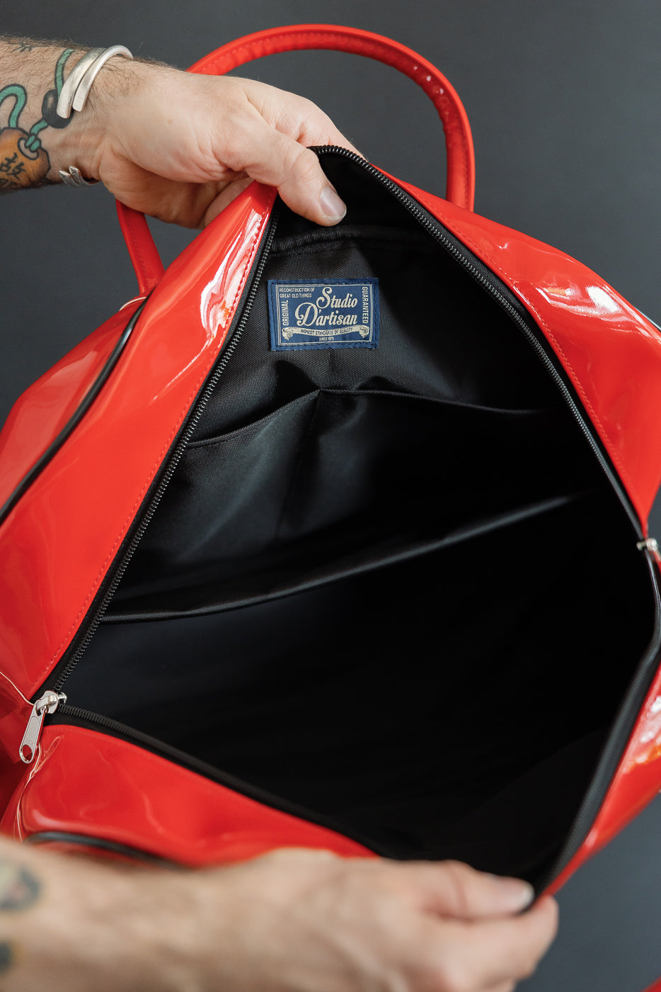 Close-up of red patent backpack interior, showing black lining, zipper, and Studio Dartisan tag, held by male model.