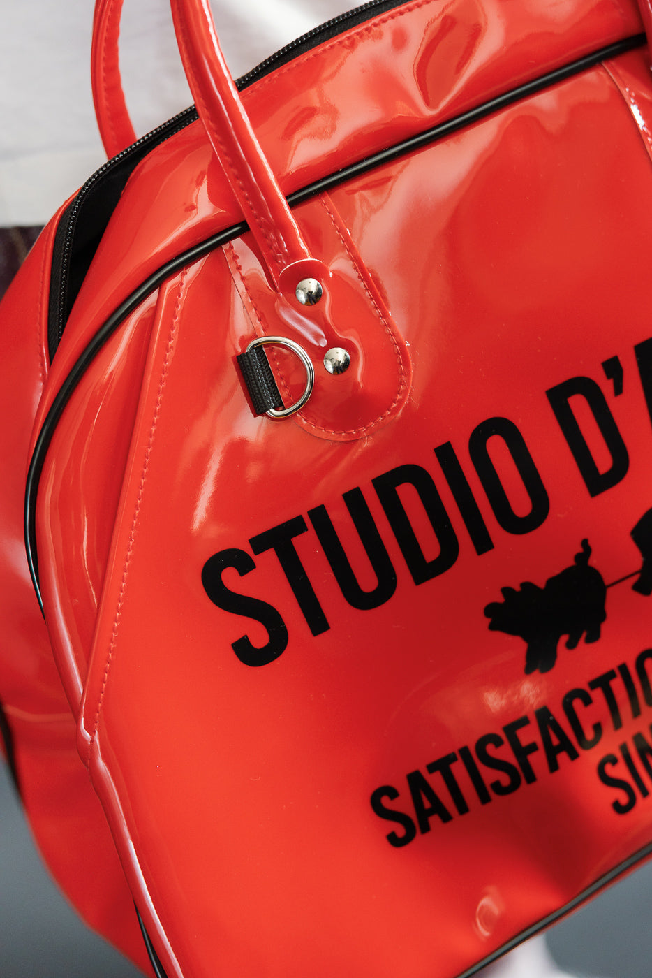 Close-up of red glossy duffel bag front, showing 'STUDIO D' print, silver hardware, and black trim details.
