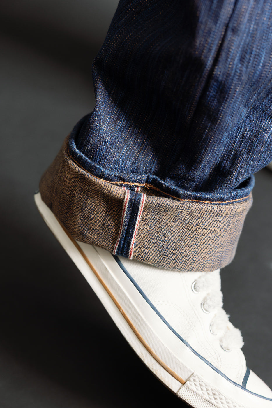 Close-up of male model’s cuffed Studio D’Artisan D1950S selvedge jeans hem, showing indigo fabric texture and selvedge ID above white sneaker.