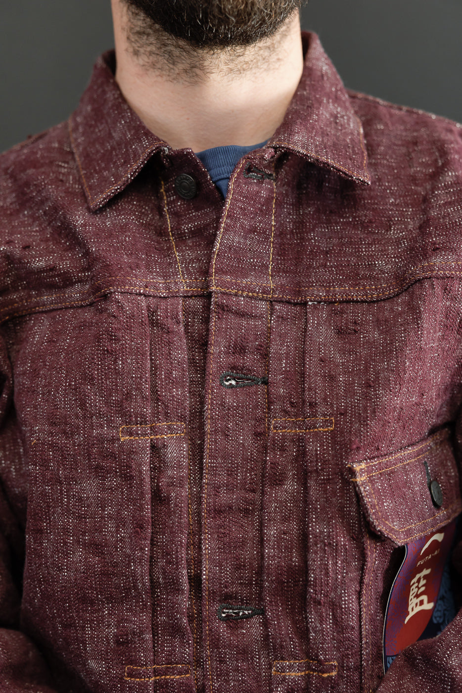 Close-up front view of male model wearing burgundy selvedge denim jacket with contrast stitching, button collar, chest pockets
