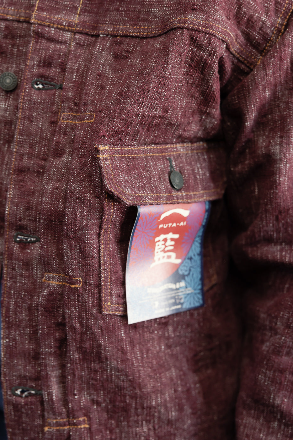 Close-up front view of burgundy selvedge denim jacket, showing chest pocket, button, orange stitching, and FUTA-AI tag detail