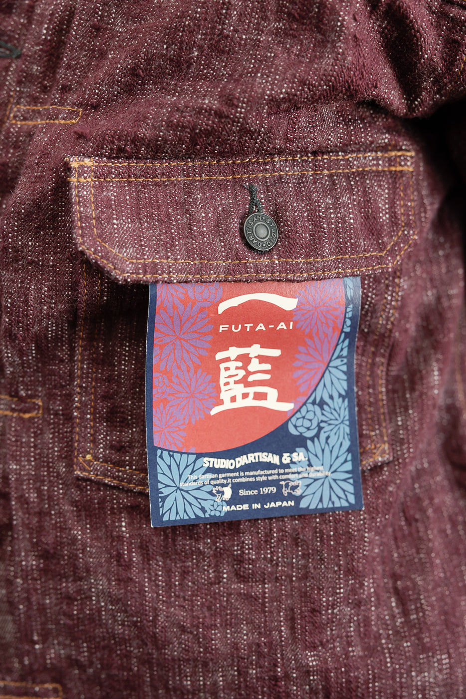 Close-up front view of burgundy selvedge denim jacket pocket with FUTA-AI tag, button, and orange stitching detail