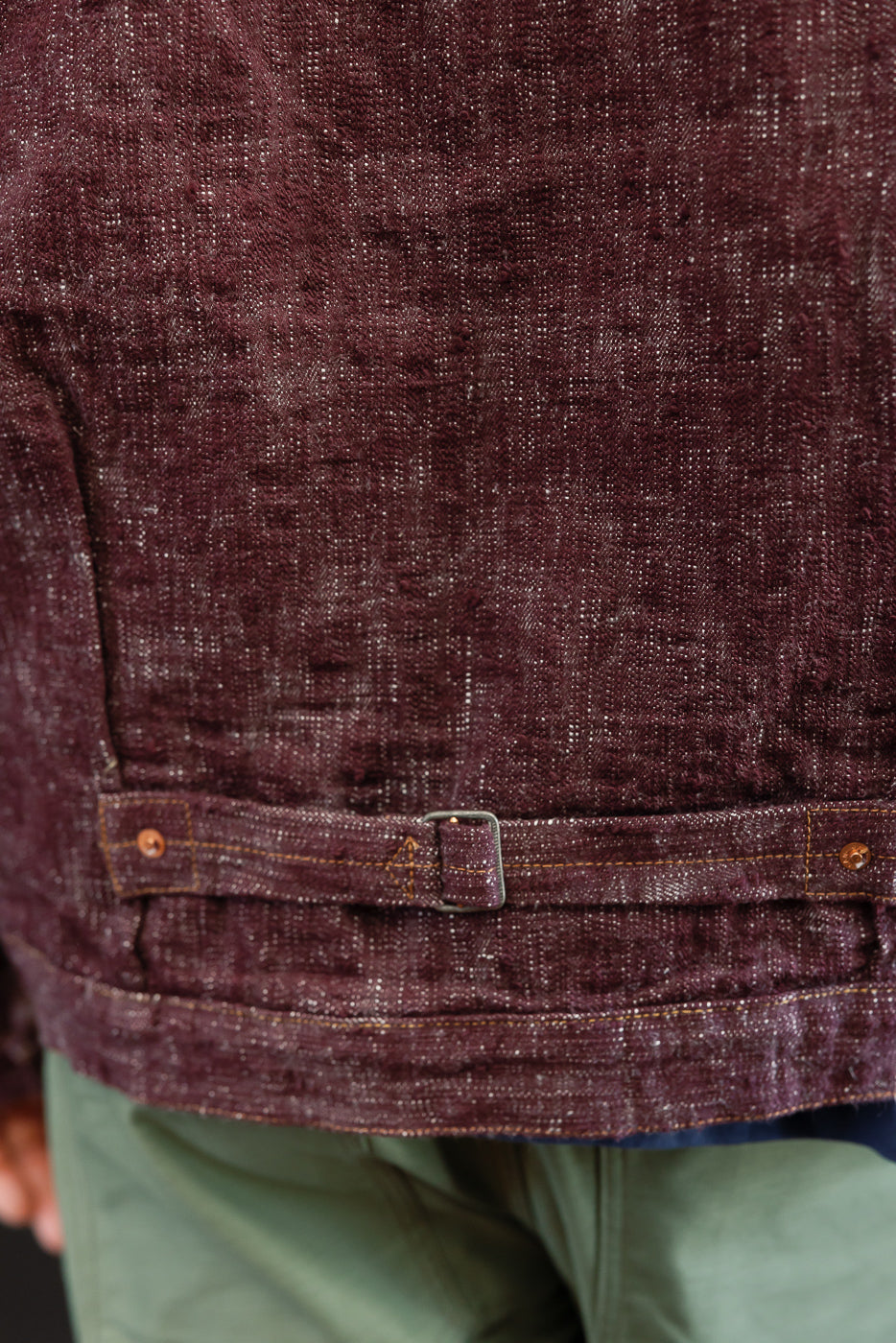 Close-up front view of maroon selvedge shirt waistband with metal buckle, contrast stitching, and fabric texture on male model.